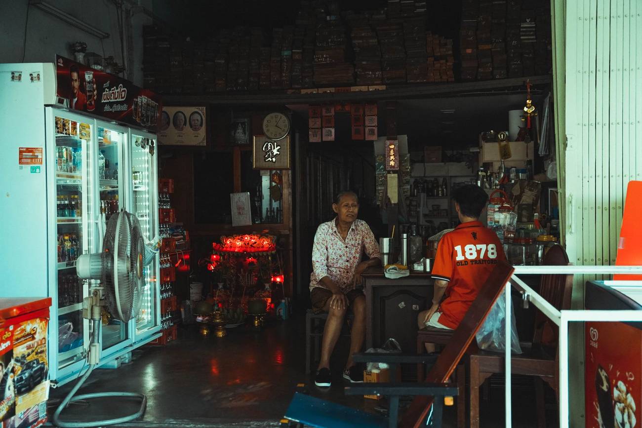 Demonstration photograph. An open shopfront in Bangkok with two men sitting inside among fridges and stacked goods.