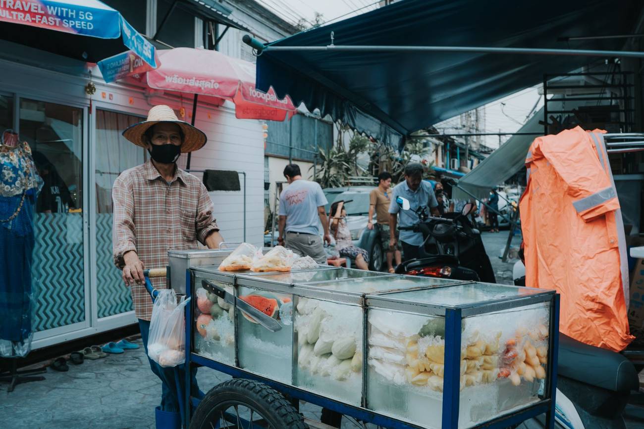 Demonstration photograph. A man in a wide hat standing behind a glass-fronted food cart in a market street.
