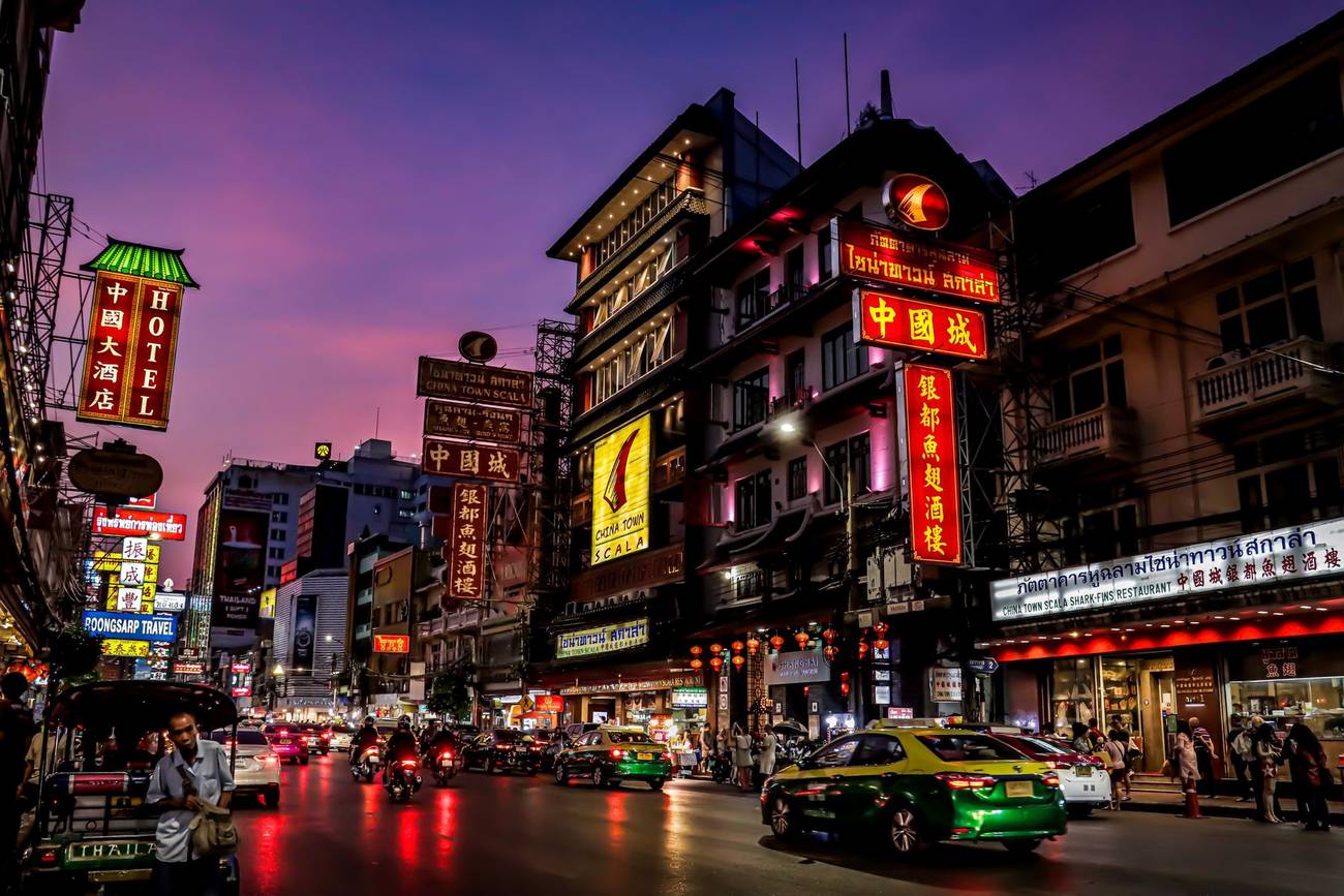 Demonstration photograph. A Bangkok street at dusk under neon signs, with a purple sky above the shophouses.